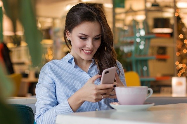 Usmiata žena v modrej košeli pije kávu a pozerá do telefónu.jpg